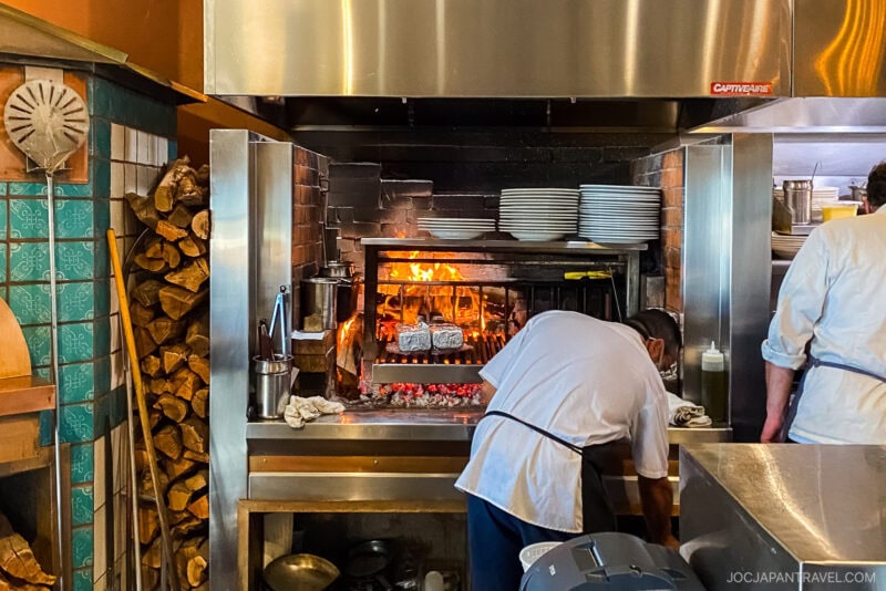 A chef tends to food cooking over an open wood fire in a California Central Coast restaurant kitchen, with neatly stacked firewood on the side and plates arranged above the cooking area.