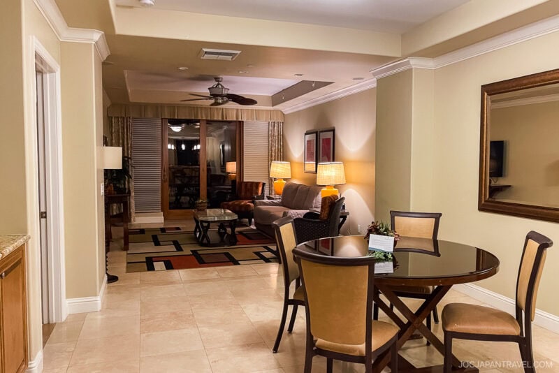 A cozy California Central Coast hotel suite with a dining table and four chairs in the foreground, a living area with a sofa, armchairs, and lamps in the background. Large windows with curtains, neutral-toned decor, and tiled floor throughout.