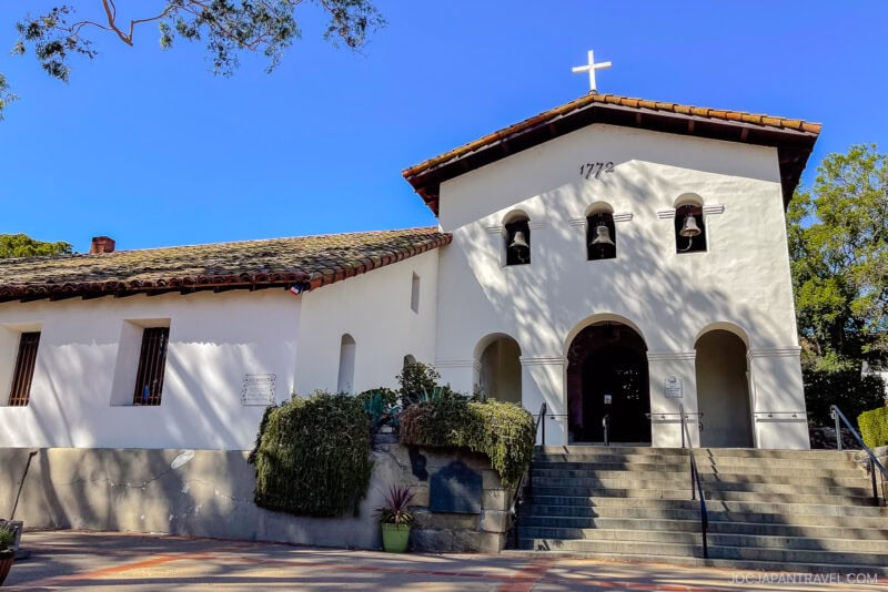 A white Spanish-style mission church with a red-tiled roof, arched entrance, three bells, and a cross on top sits under a clear California Central Coast sky. Stairs lead up to the entrance, plants line the front, and "1772" is above the door.