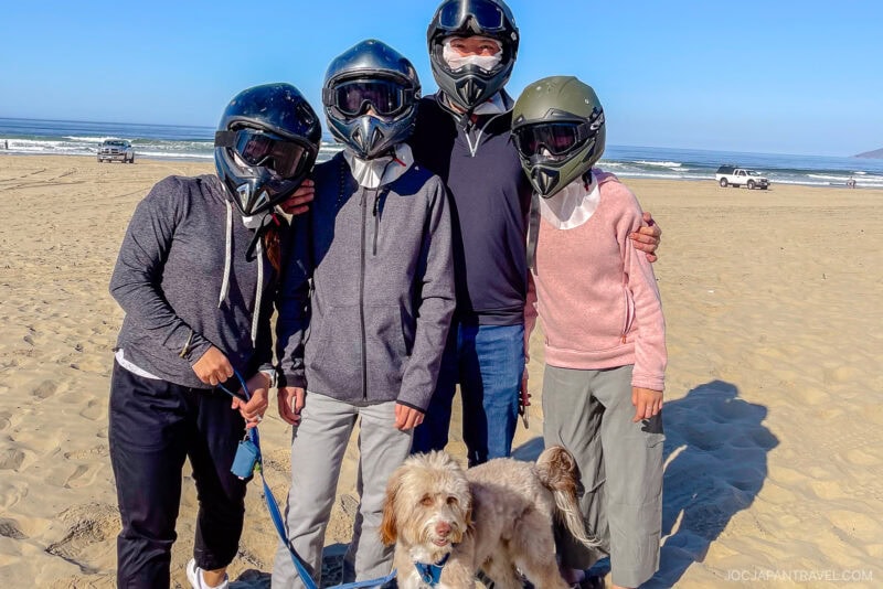 Four people wearing helmets stand together on a sandy California Central Coast beach with the ocean in the background. A small, light brown dog on a leash stands in front of them. Vehicles and waves are visible in the distance.