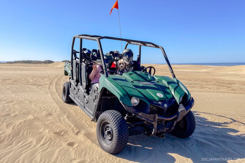 A green off-road vehicle with several people inside, wearing helmets and sunglasses, is parked on sandy desert terrain under a clear blue sky on the California Central Coast. An orange flag is attached to the vehicle.