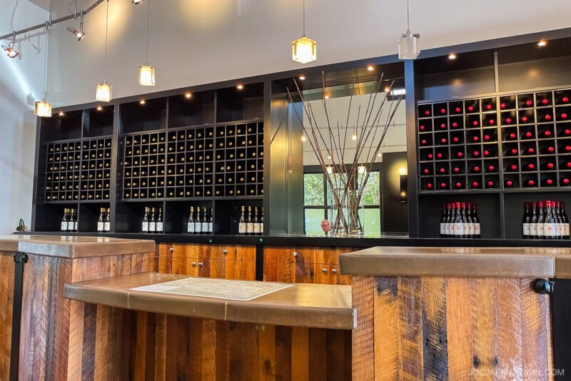 Modern wine tasting room with wooden counters, a California Central Coast map on the counter, and a black wall display filled with wine bottles and branches in a vase as decor. Hanging lights illuminate the clean, organized space.