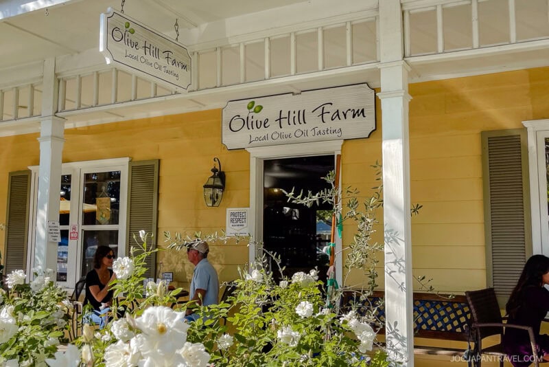 People stand outside Olive Hill Farm, a yellow building with white trim on the California Central Coast offering local olive oil tasting. White flowers and greenery frame the porch, while a sign hangs above the entrance.