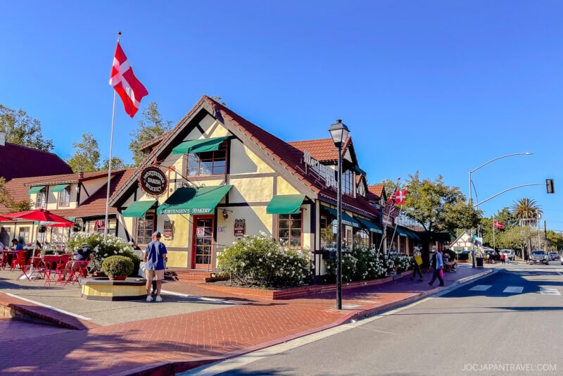 A Danish-style bakery on the California Central Coast features a red-and-white flag, green awnings, and outdoor seating on a sunny street corner. A few people stroll by under the clear blue sky.