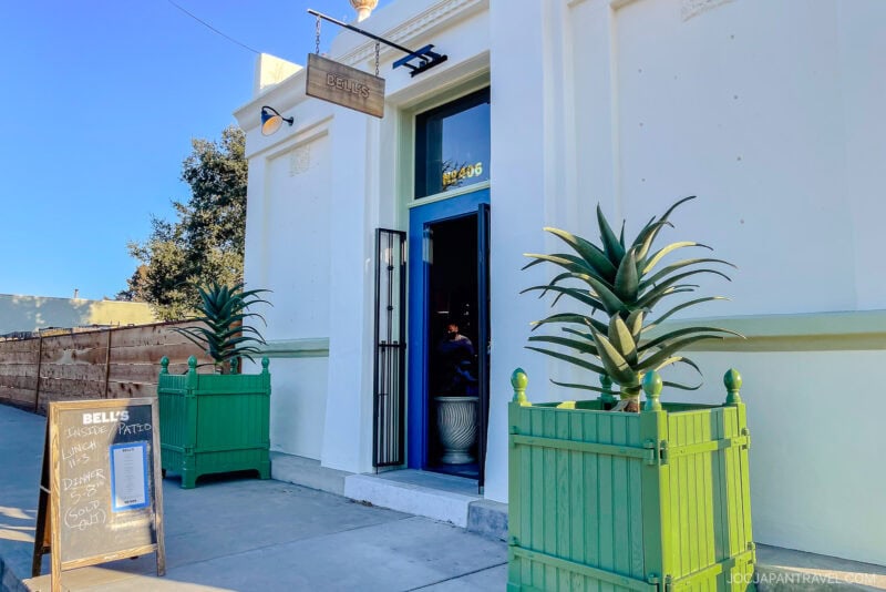 A white building with a blue door and a wooden sign reading “Belli’s” evokes California Central Coast charm. Two large green planters with agave plants flank the entrance, and a chalkboard menu stands on the sidewalk in front.