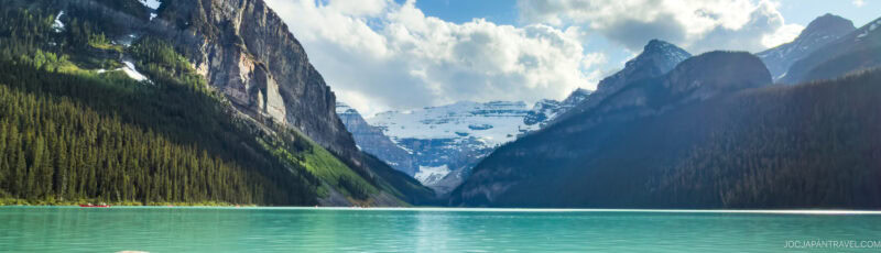 A turquoise lake surrounded by dense evergreen forests and tall, snow-capped mountains under a partly cloudy sky. The landscape is tranquil and scenic, with dramatic peaks reflecting in the clear water.