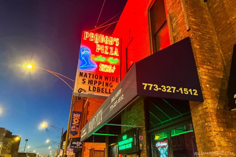 Neon signs for Pequod’s Pizza and nationwide shipping glow above a brick building at dusk in Chicago, showcasing the iconic Chicago food scene amid streetlights and a deep blue sky in the background.