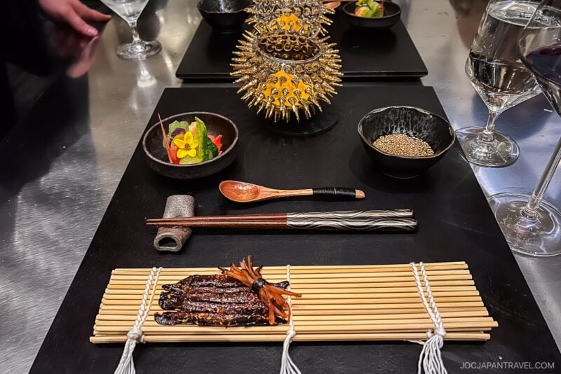 A gourmet Japanese meal in Chicago Food style, featuring grilled seafood on a bamboo mat, a ceramic pipe, chopsticks, wooden spoon, small bowls of garnish, and an ornate spiky sea urchin shell on a black tray with a glass of water.