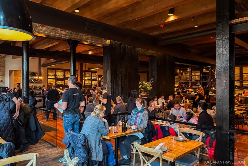 A bustling, warmly lit Chicago Food restaurant with wooden beams, diners seated at tables, and servers attending to guests. The bar area is visible in the background, creating a lively and welcoming atmosphere.