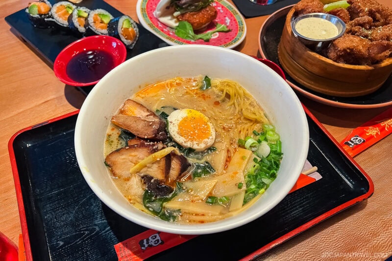A bowl of ramen with sliced pork, half a boiled egg, bamboo shoots, green onions, and broth is served on a tray—classic Chicago Food—alongside sushi rolls, fried chicken with dip, and a small plate of appetizers.