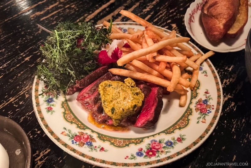 A floral plate with sliced steak topped with herb butter, French fries, and a small salad on a dark wooden table—a classic Chicago food presentation. A piece of bread is partially visible on a separate plate in the background.