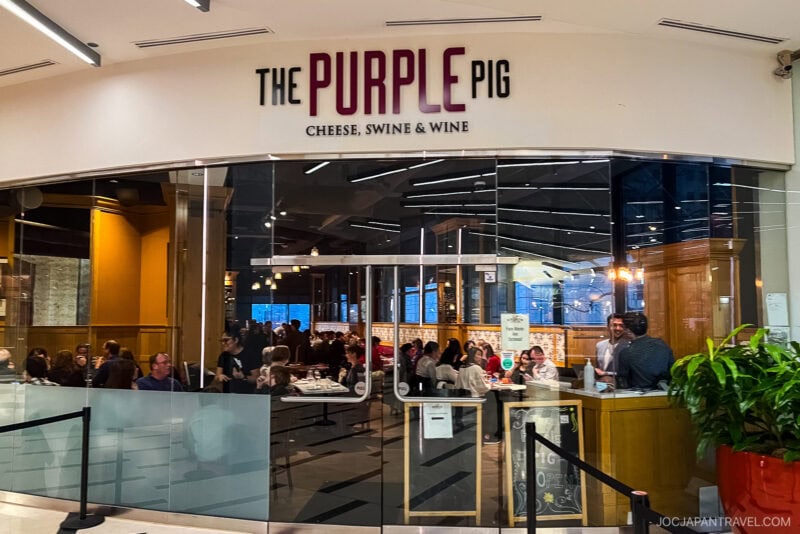 The entrance to The Purple Pig restaurant in Chicago, with glass doors and a large sign reading The Purple Pig: Cheese, Swine & Wine. Diners savoring Chicago food are visible inside, seated at tables, while staff welcomes guests near the entrance.