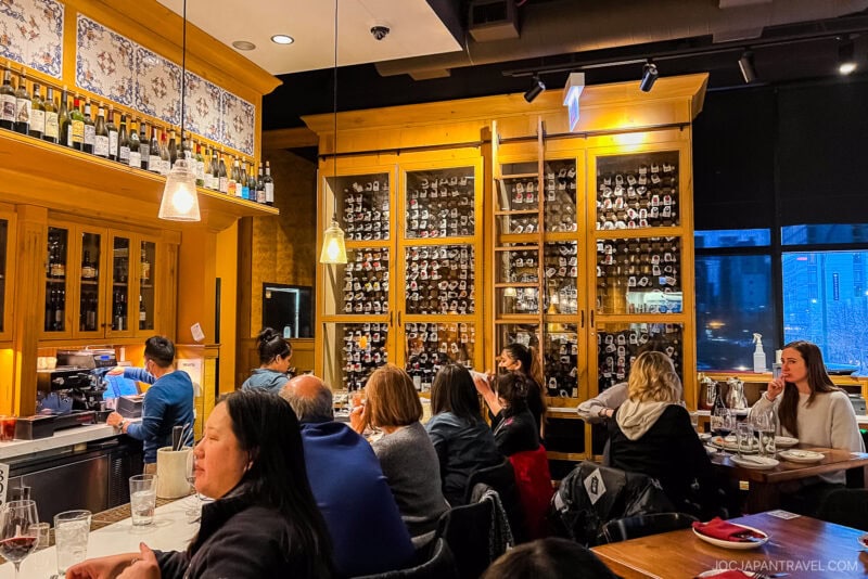 A cozy Chicago food restaurant scene with diners seated at tables and a bar. Shelves behind the counter display wine bottles and glassware, while warm lighting and large windows create an inviting atmosphere.