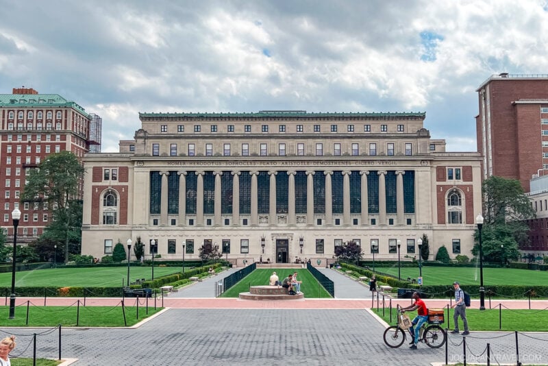 A large neoclassical university building with tall columns, surrounded by green lawns and people walking or riding bikes in the foreground under a partly cloudy sky—an iconic scene on any Northeast College Tour.