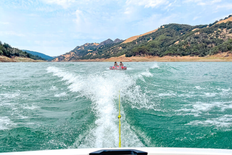 A view from a boat looking back at two people tubing on a Sonoma lake, with a wake trailing behind and green hills in the background under a partly cloudy sky.