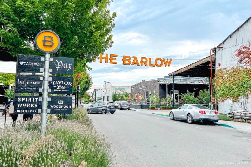 A street view of The Barlow in Sonoma, a modern outdoor marketplace with shops, restaurants, and signs for local businesses. Cars are parked along the road, trees and greenery line the sidewalk, and the sky is partly cloudy.