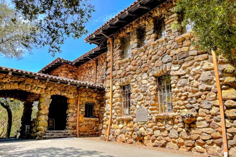 A historic stone building with a tiled roof and arched entryway stands under the sunny Sonoma sky, surrounded by green trees. A metal plaque is mounted between two windows on the stone wall.