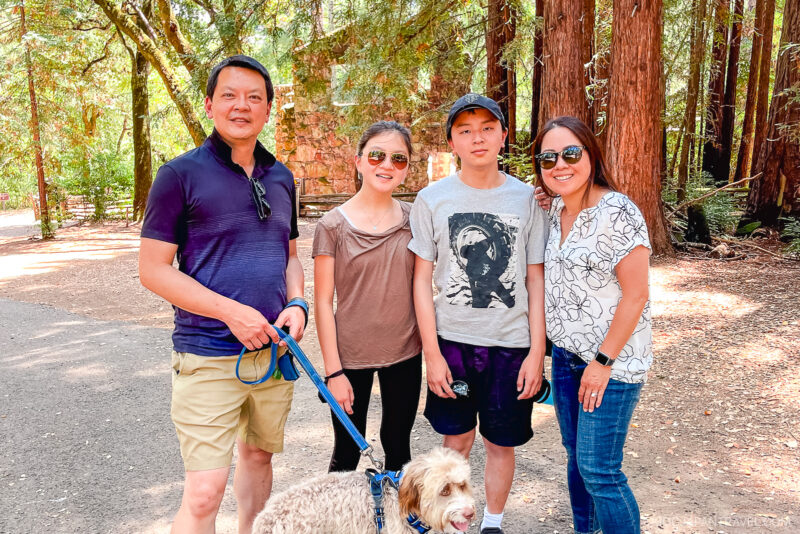 A family of four poses outdoors with their dog on a leash along a sunny Sonoma dirt path surrounded by tall trees. Everyone is smiling at the camera, enjoying the beautiful day.