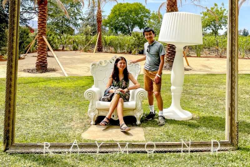 Two people pose in a Sonoma outdoor park framed by a giant picture frame. One sits on a decorative white chair while the other stands beside a large white lamp. Palm trees and greenery fill the background. The word RAYMOND appears below.