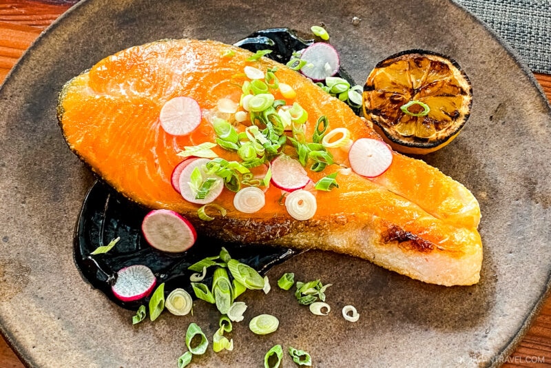 A cooked salmon steak garnished with sliced radishes and green onions, served on a brown plate with a charred lemon half and a dark Sonoma-inspired sauce underneath.