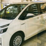 Driving in Japan – Tips for Travelers A white HYBRID minivan, highlighted in this Japan driving guide, is parked indoors near a garage or service area, with reflections visible on its windows and body.