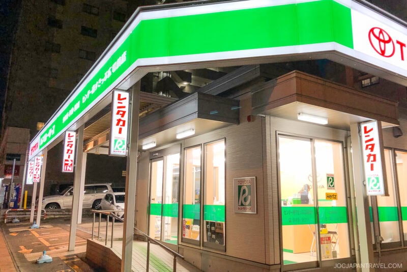 A brightly lit Toyota Rent a Car and Hertz rental office at night in Japan, with green and white signage and large windows showing the interior counter—an ideal stop for travelers following a japan driving guide.
