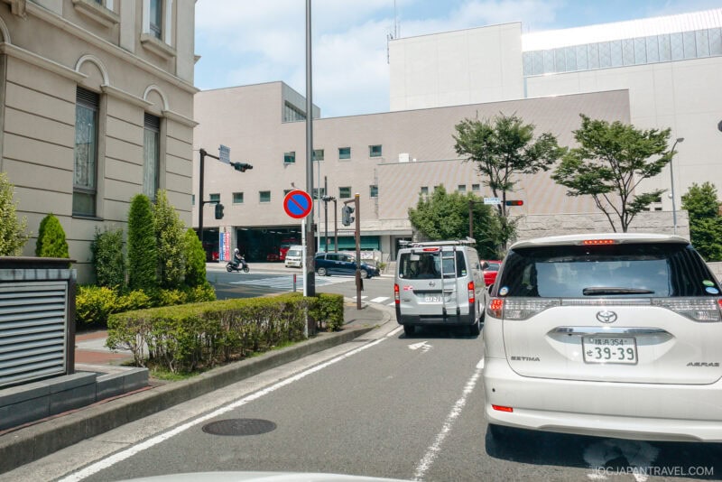 Cars wait at a traffic light on a city street bordered by buildings, shrubs, and trees. A blue and red no parking sign is visible, the sky partly cloudy—a typical scene from any japan driving guide, complete with Japanese license plates.