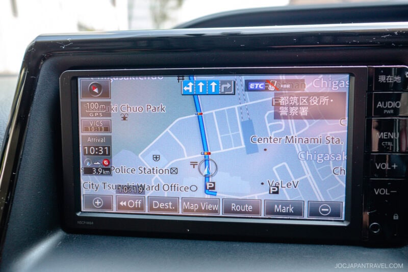 A car GPS navigation screen displays a map with a blue route line, local landmarks, Japanese text, and estimated arrival time of 10:31. Several control buttons are visible below the map, making it ideal for any japan driving guide.