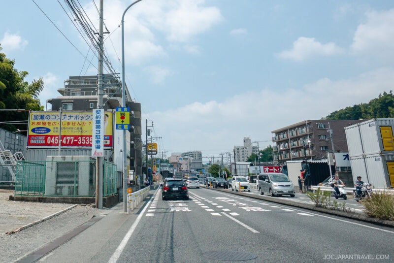A street scene in Japan with cars driving, buildings and apartments on both sides, Japanese signs, utility poles, and pedestrians on the sidewalk under a partly cloudy sky—a perfect setting for a japan driving guide.
