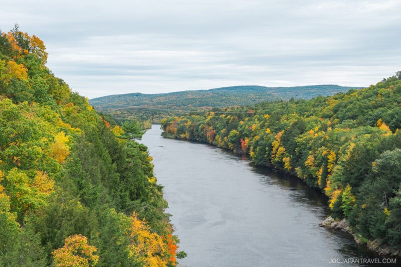 A wide river winds through a lush New England forest with trees showing green, yellow, and orange autumn foliage, beneath a cloudy sky with distant hills in the background.