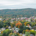 Fall Foliage & Freedom Trail – 4 Days in New England A New England town with houses and buildings surrounded by trees in vibrant autumn foliage, set against rolling hills covered in colorful fall leaves under a cloudy sky.