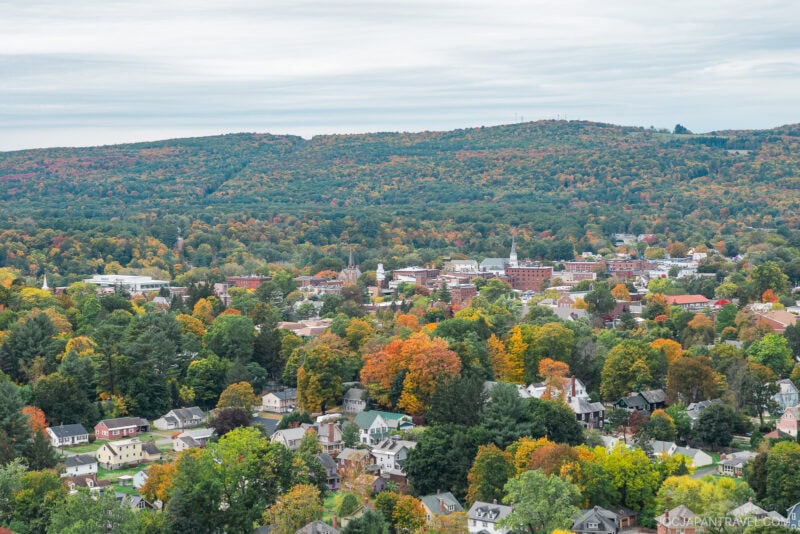 A New England town with houses and buildings surrounded by trees in vibrant autumn foliage, set against rolling hills covered in colorful fall leaves under a cloudy sky.