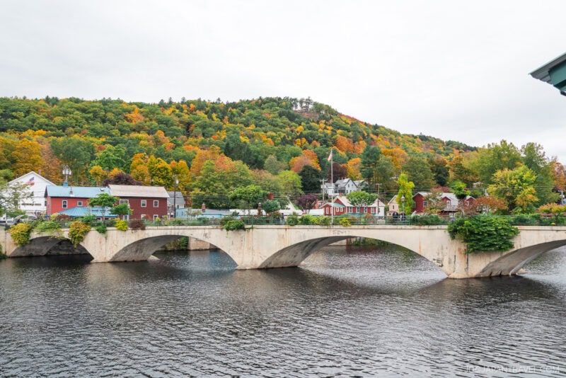 A stone bridge with arches crosses a river in New England; charming houses and colorful autumn trees line the riverbank, with a forested hill rising in the background under an overcast sky.