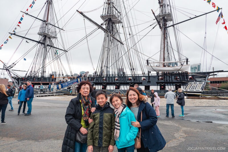 Four people smile and pose together in front of a historic New England sailing ship with tall masts and rigging. Other visitors walk nearby under a cloudy sky, while city buildings stand visible in the background.