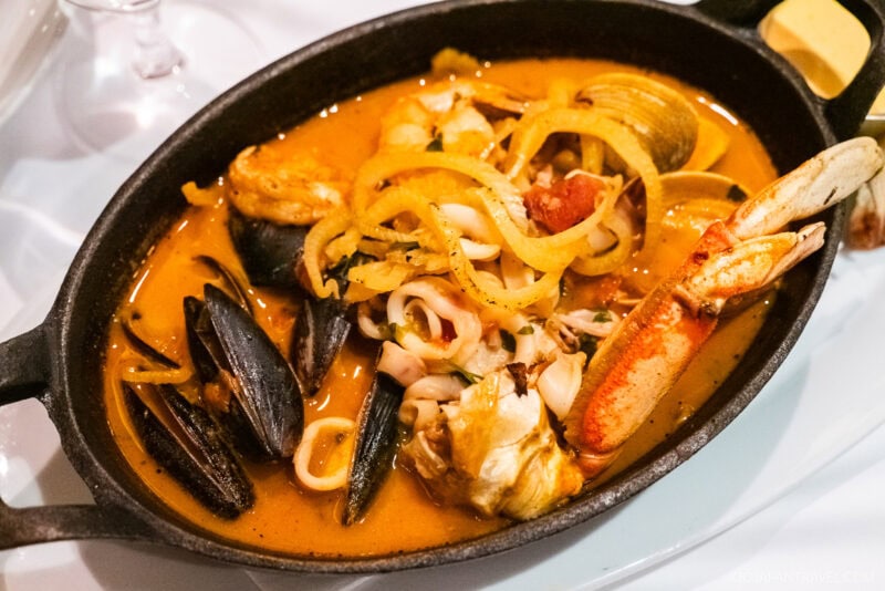 A New England seafood stew in a black dish featuring mussels, clams, crab, squid, and fish in a rich orange broth, garnished with sliced onions.