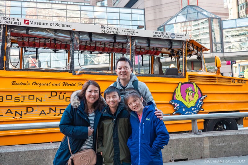 A smiling family of four poses in front of a bright yellow Boston Duck Tours vehicle outside the Museum of Science, ready to explore New England. City buildings rise in the background alongside signage for tickets and departures.