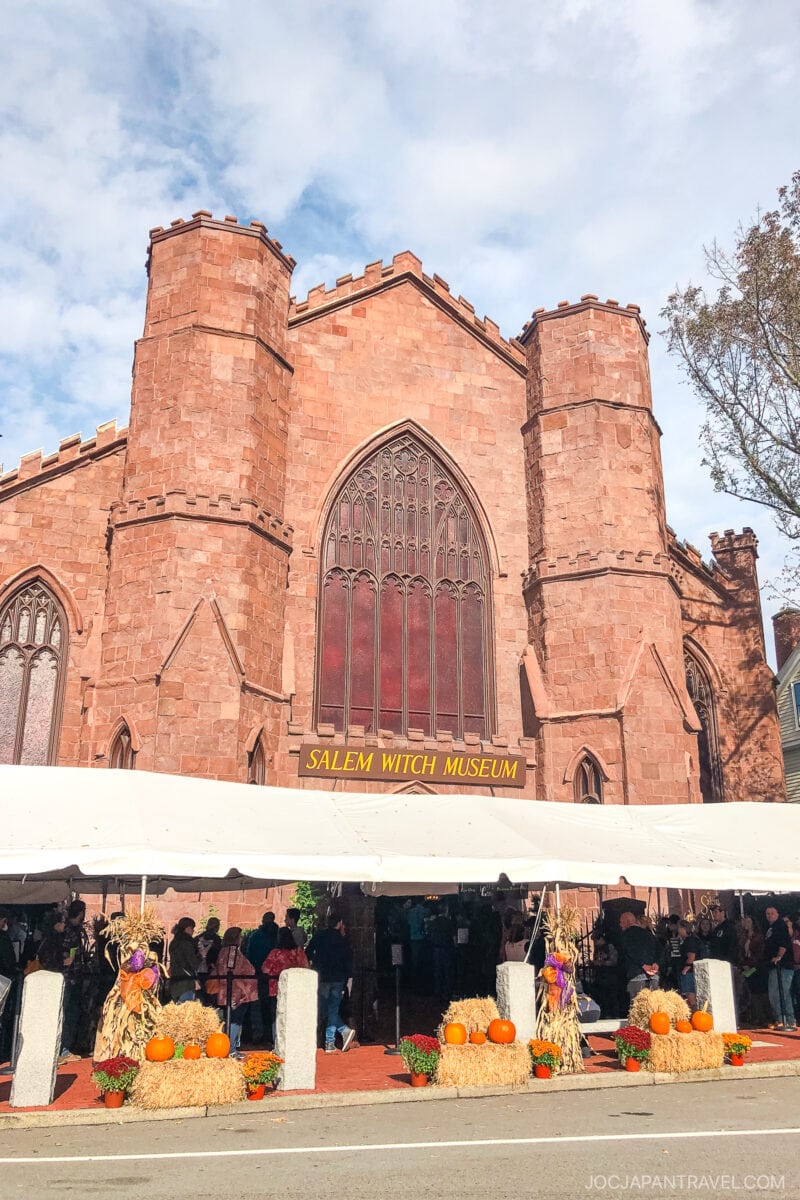A large red-brick Gothic-style building labeled Salem Witch Museum sits behind a white tent decorated with pumpkins, hay bales, and autumn flowers—capturing classic New England charm as people gather under the tent on a sunny day.