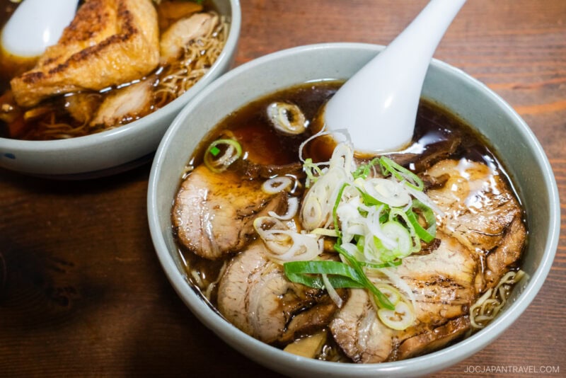 Two bowls of ramen, a signature food in Takayama, with sliced pork, green onions, and dark broth sit on a wooden table. One bowl features fried tofu. White ceramic soup spoons rest inside each bowl.