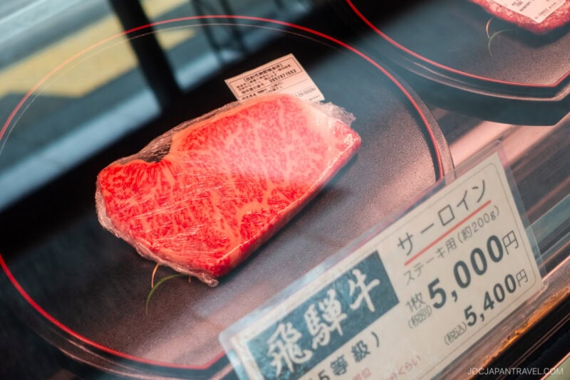 A piece of marbled Wagyu steak, wrapped in plastic, is displayed on a black plate with a 5,000 yen price tag in a store showcase—highlighting premium food in Takayama. Japanese labels are visible beside the steak.