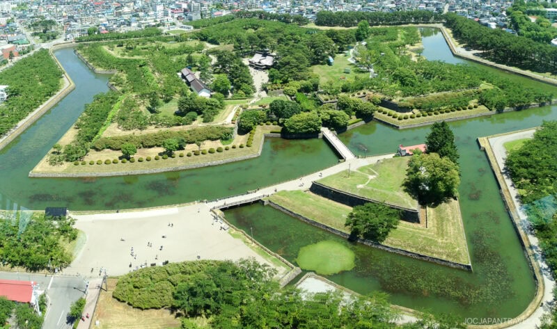Aerial view of Fort Goryokaku, a star-shaped fort surrounded by a moat and green trees, with walkways, bridges, and people exploring the historic Hakodate site in Japan.