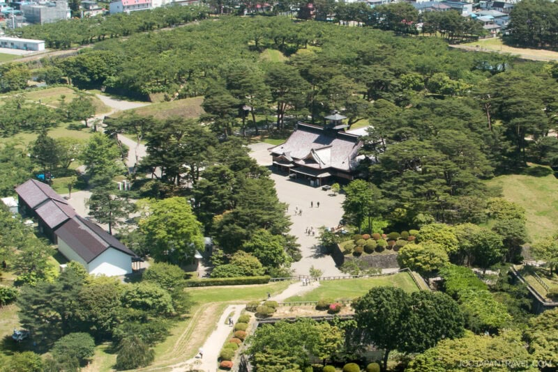 Aerial view of Fort Goryokaku, a historic Japanese building surrounded by dense green trees and gardens, with walkways and people scattered around the complex. Smaller white buildings are nearby, and a city is visible in the background.