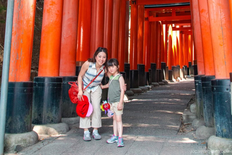 A woman and a young girl pose together in front of a path lined with vibrant red torii gates at Fushimi Inari Shrine in Japan on a sunny day.