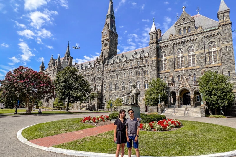 Two people stand smiling in front of a large historic stone building with a clock tower, surrounded by flowers and greenery under a bright blue sky. An airplane soars above, marking an exciting stop on their Northeast College Tour.