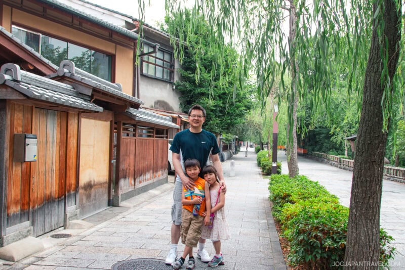 A man stands on a stone-paved street in Gion, Japan, smiling with two young children before him. Traditional wooden buildings and weeping willow trees line the quiet street, capturing the charm of this historic district.