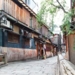 Gion (祇園) – Kyoto’s Historic Geisha District A narrow stone-paved street in Gion lined with traditional wooden buildings, lanterns, and signs. A person in a kimono walks in the distance as green trees hang over the peaceful scene.