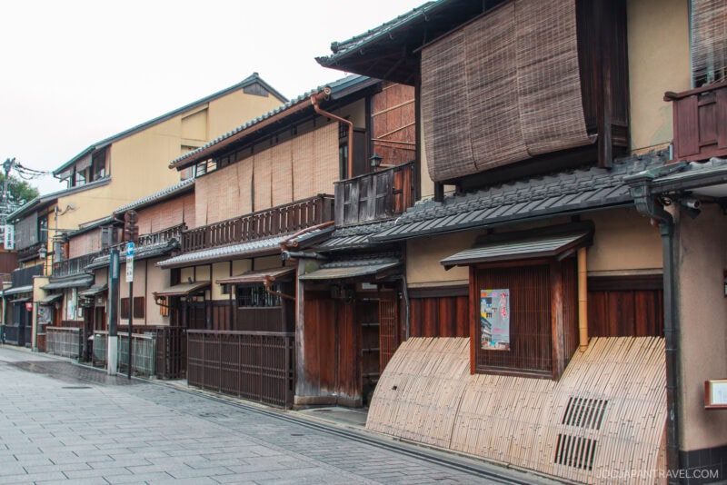 A quiet street in Japan’s Gion district lined with traditional wooden buildings featuring sliding doors, bamboo shades, and tiled roofs, with no people visible.