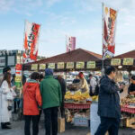 People gather at outdoor market stalls in Hachinohe, browsing and buying snacks and produce. The colorful banners and signs feature Japanese text. Several customers wear jackets beneath the partly cloudy sky.
