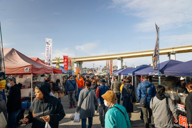 People walk through a busy outdoor market in Hachinohe, lined with tents and stalls on both sides. Colorful banners and signs brighten the scene, while an overpass stretches across the background under a partly cloudy sky.