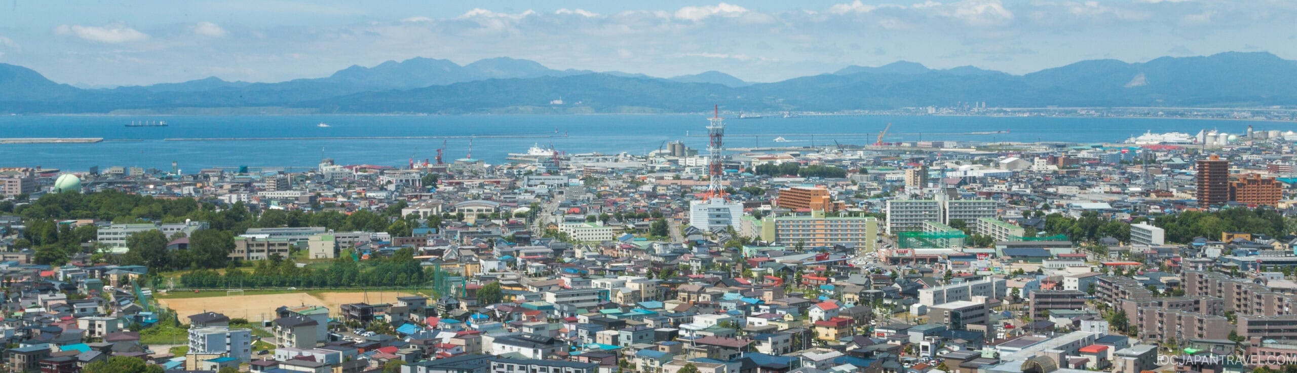 A panoramic view of a coastal city with dense buildings, a harbor with ships, and a backdrop of blue mountains under a partly cloudy sky.