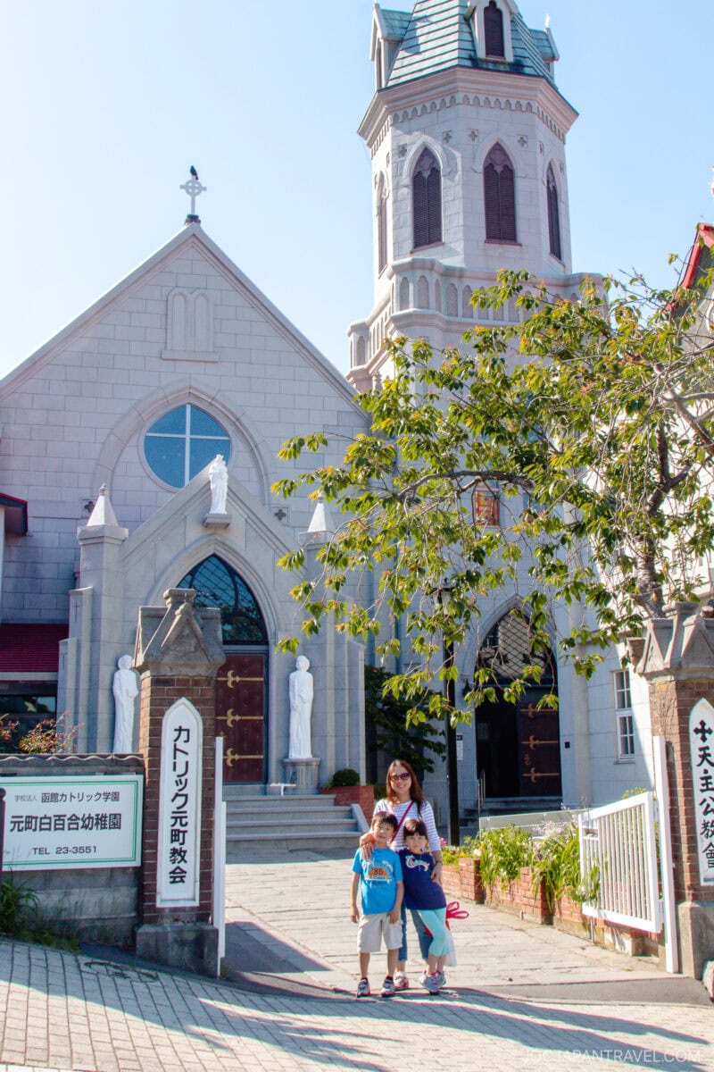 A woman and two children stand smiling in front of a stone church with arched windows, a clock tower, and statues near the entrance in Hakodate Motomachi District. A leafy tree partially shades the path leading to the church.